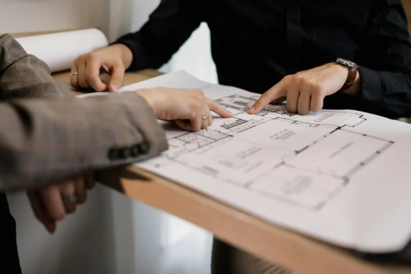 Two people point at sections of a printed floor plan or architectural blueprint on a desk, discussing details.