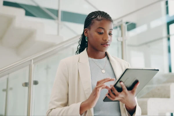 A woman in business attire uses a tablet while standing near a staircase in a modern, brightly lit office building.