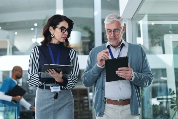 Two business professionals with lanyards review information on digital tablets while walking through a modern office environment.