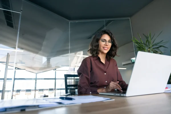 A woman wearing glasses and a brown shirt sits at a desk in a modern office, working on a laptop. Papers and a pen are visible in the foreground.