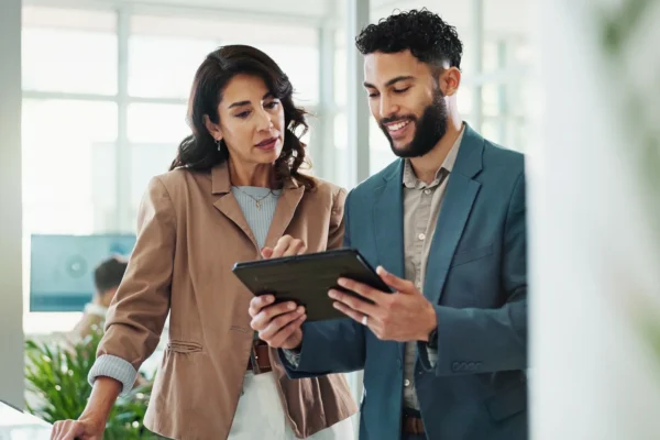 Two people in business attire stand together in an office, looking at and discussing information on a tablet device.
