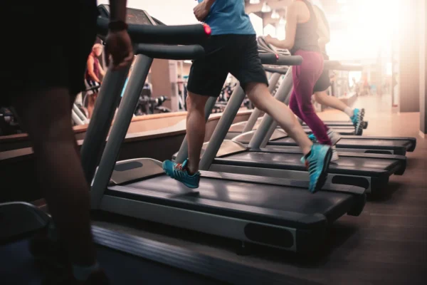 People are walking or running on treadmills in a gym, with focus on their legs and feet. The room is well-lit and several treadmills are in use.