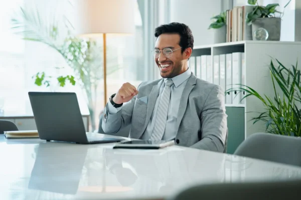 A man in a gray suit sits at a desk, smiling and raising his fist in front of an open laptop in a modern office setting with plants and shelves in the background.