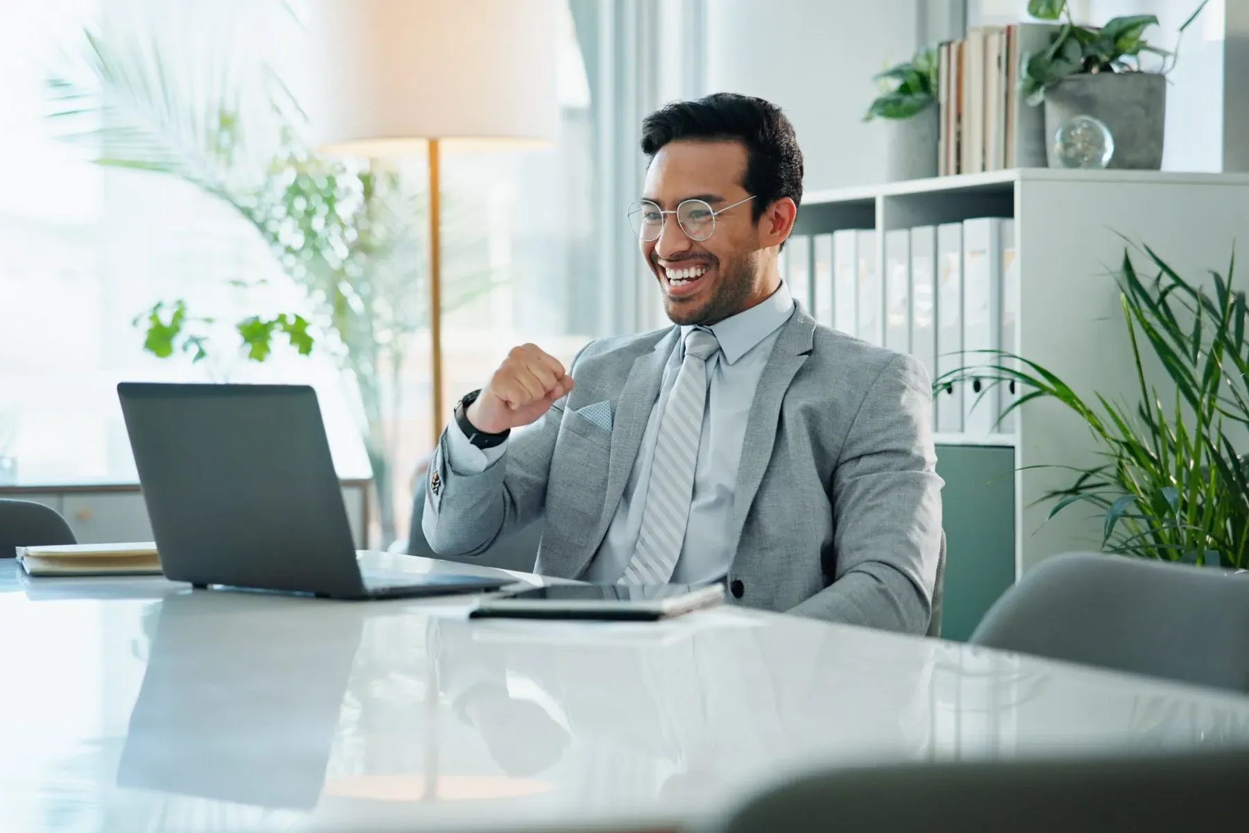 A man in a gray suit sits at a desk, smiling and raising his fist in front of an open laptop in a modern office setting with plants and shelves in the background.