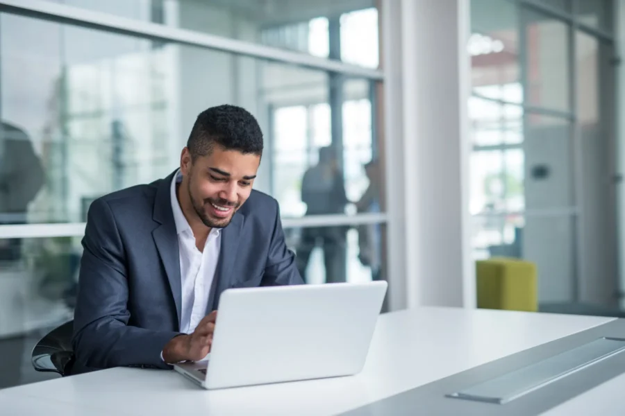 A man in a suit sits at a desk working on a laptop in a modern office with glass walls and people in the background.