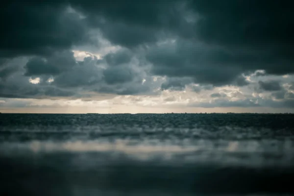 Dark clouds cover the sky over a calm sea, with a faint horizon line visible in the distance and diffused sunlight breaking through.