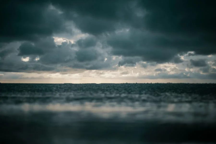 Dark clouds cover the sky over a calm sea, with a faint horizon line visible in the distance and diffused sunlight breaking through.