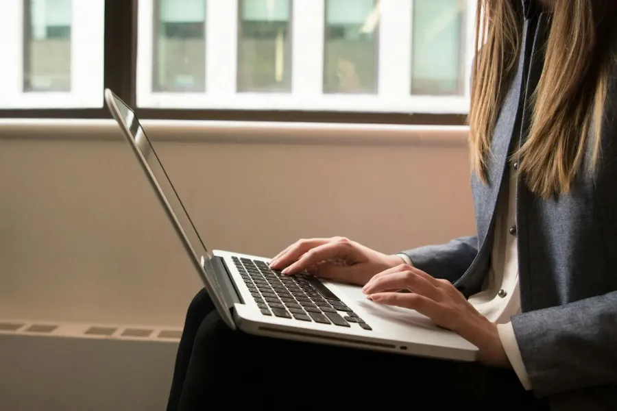 Person in business attire typing on a laptop while seated indoors near a window with buildings visible outside.