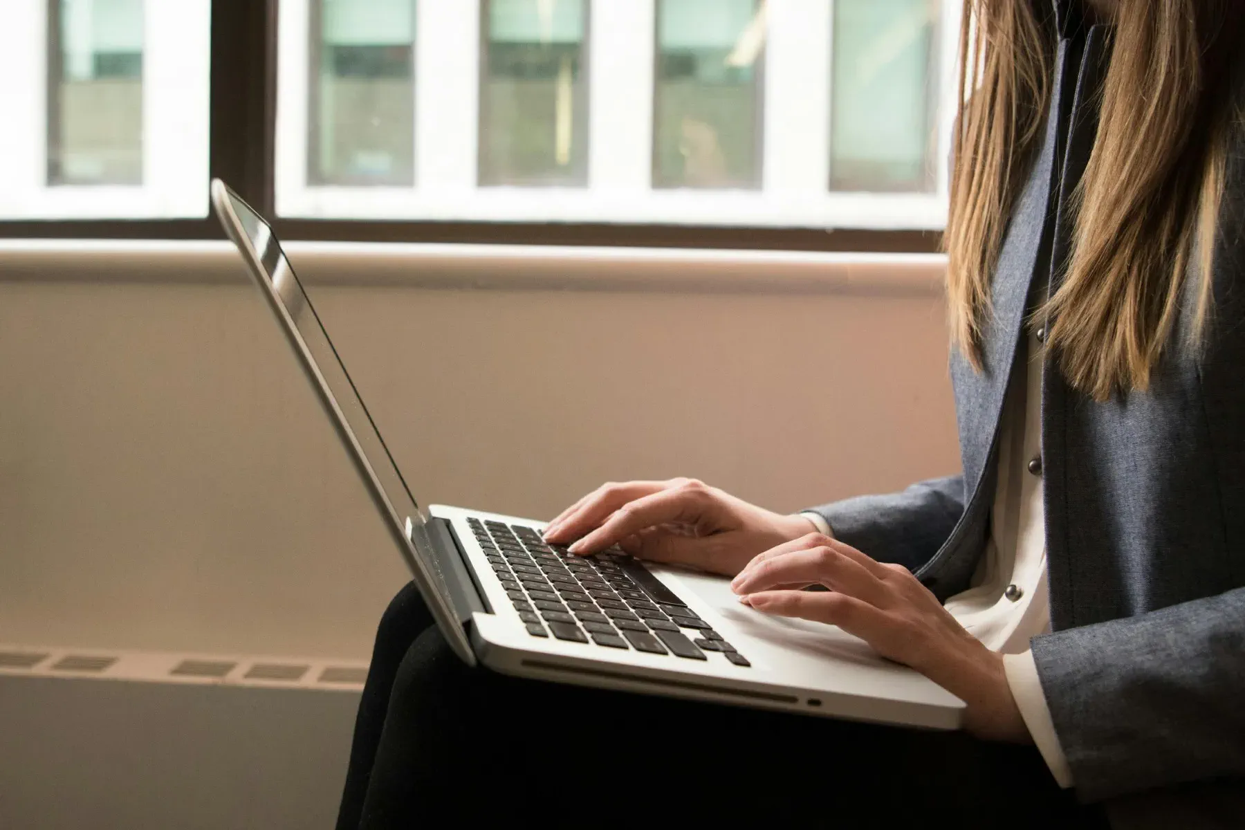 Person in business attire typing on a laptop while seated indoors near a window with buildings visible outside.