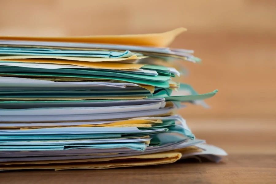 A close-up of a stack of assorted papers and folders on a wooden surface, with documents in various colors and slightly uneven edges.