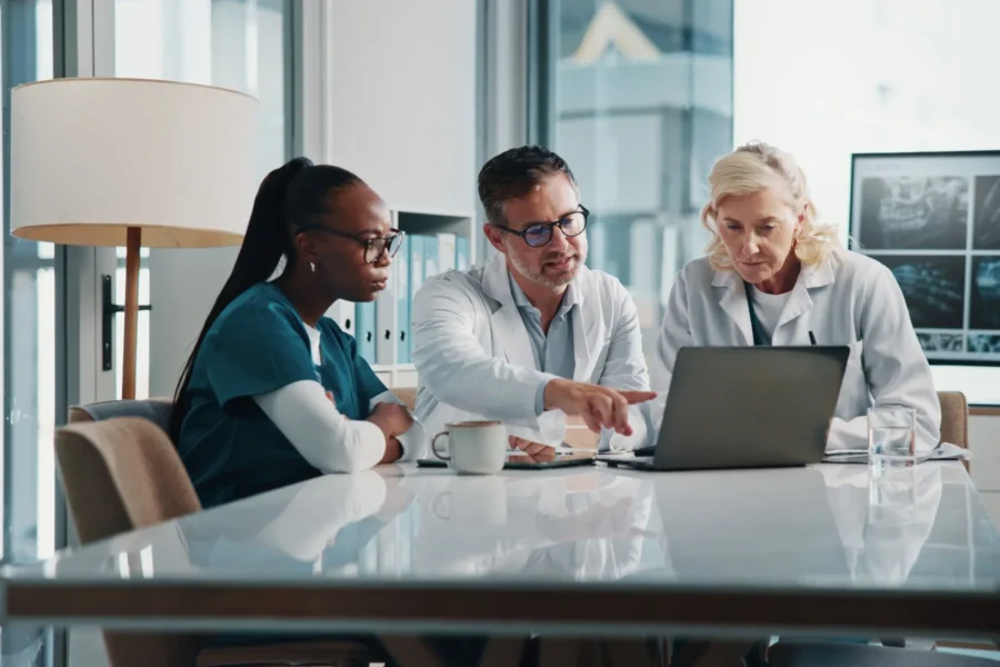 Three medical professionals review information on a laptop at a conference table, with medical images visible on a screen in the background.