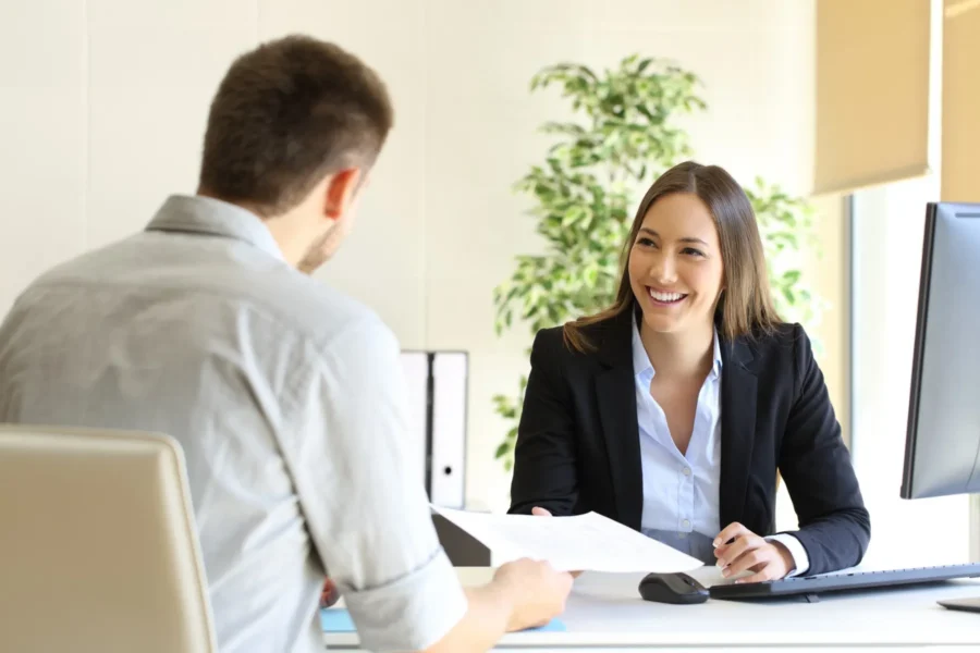 A woman in business attire smiles while talking to a man across a desk in an office setting, with a computer monitor and documents on the desk.