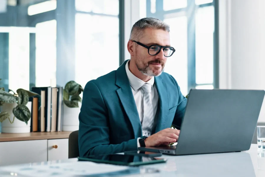 A man in a blue suit and glasses works on a laptop at a desk in a modern office with plants and shelves in the background.