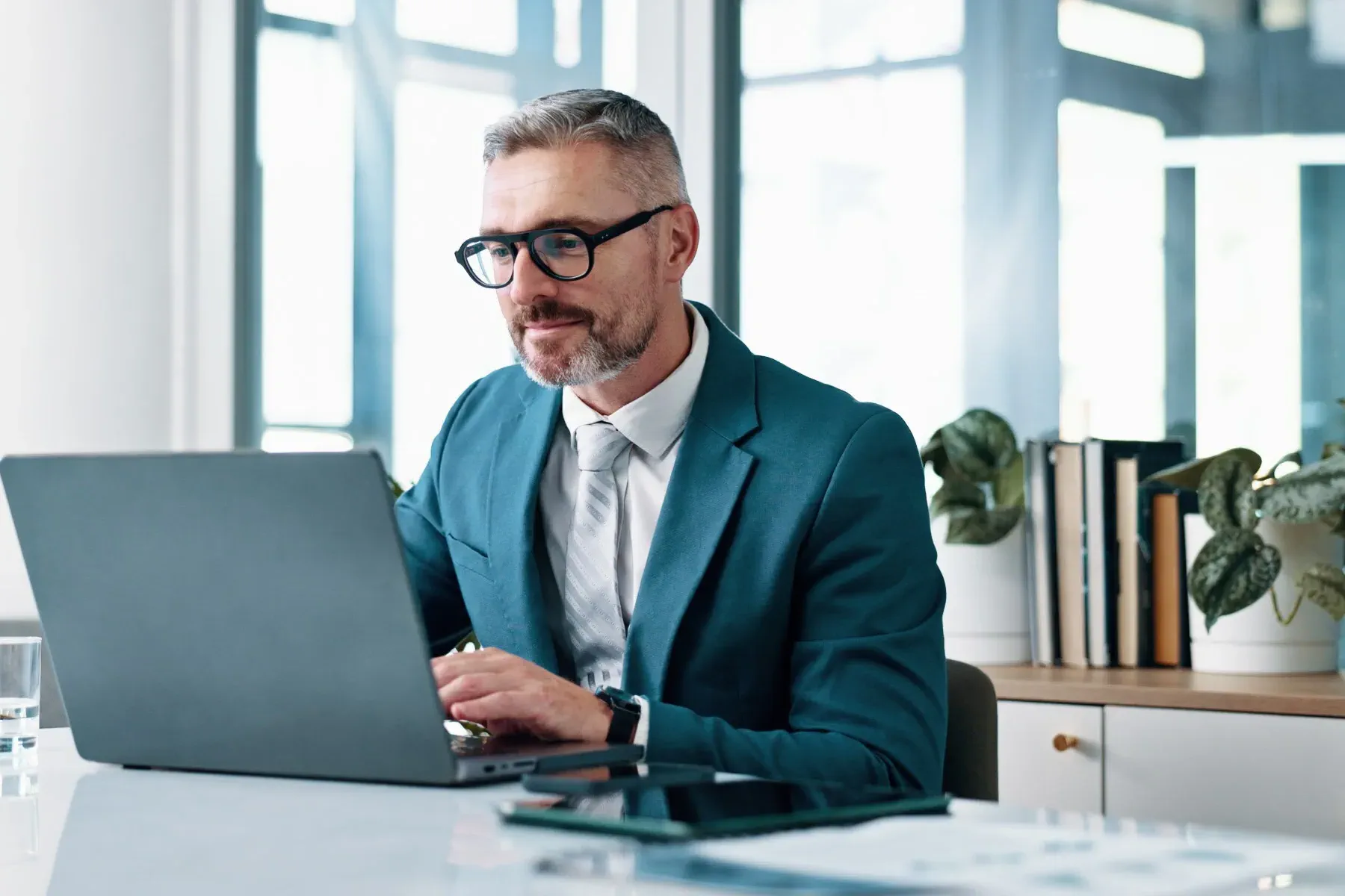 A man wearing glasses and a teal suit works on a laptop at a desk in a modern office with plants and books in the background.
