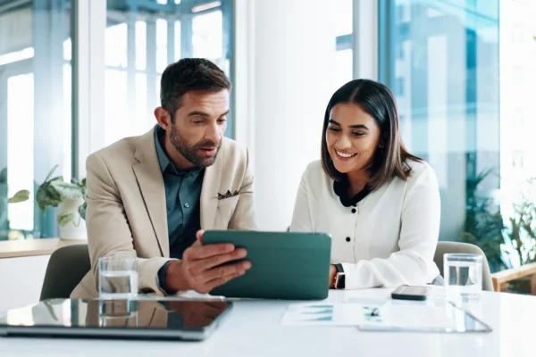 Two people sit at a desk in an office, looking at a tablet together. Papers, a laptop, and glasses of water are on the table.