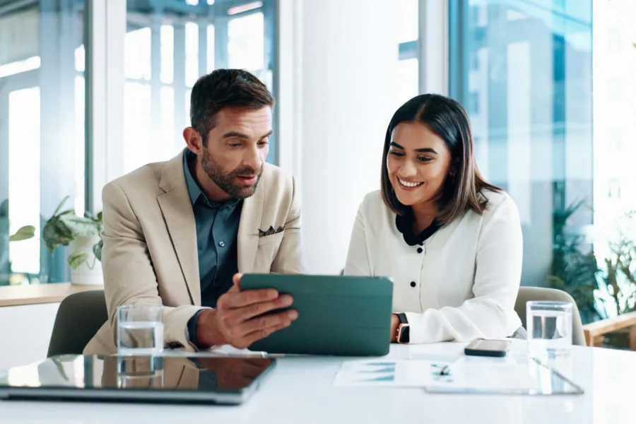 Two people sit at a desk in an office, looking at a tablet together. Papers, a laptop, and glasses of water are on the table.