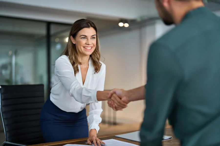 Woman in business attire smiling and shaking hands with a man across a desk in a modern office setting.
