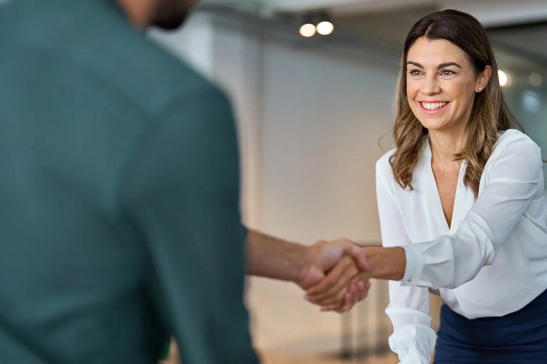 Woman in white blouse smiles while shaking hands with an unseen man in a modern office setting.