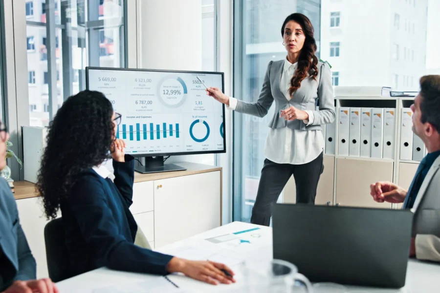 A woman in business attire presents data charts on a large screen to colleagues seated around a conference table in a modern office.
