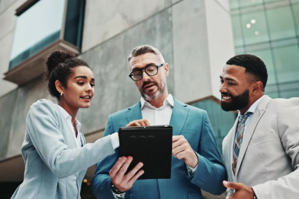 Three business professionals in suits looking at a tablet together outdoors.