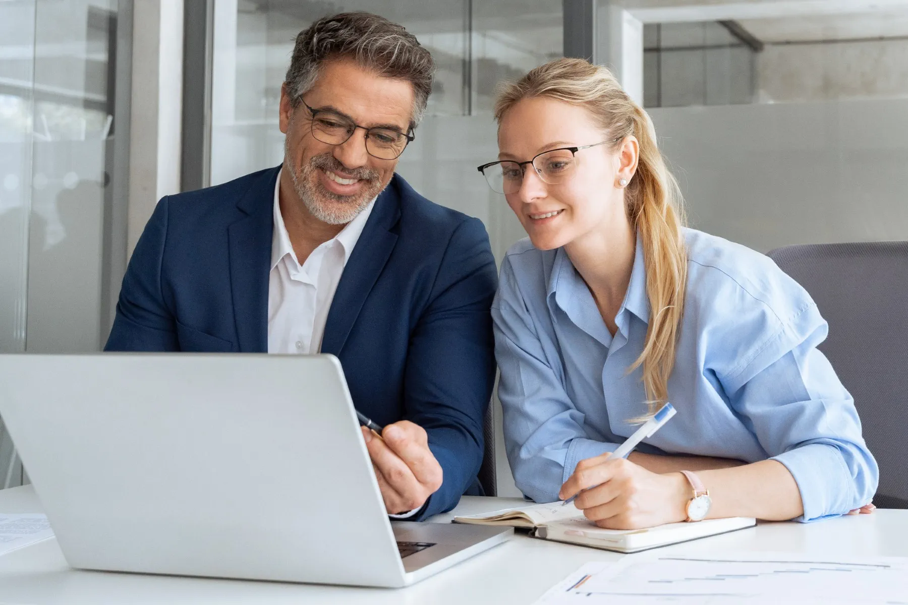 Two people in business attire sit at a desk, looking at a laptop screen. One holds a pen and notebook, and they appear to be discussing work or collaborating on a project.