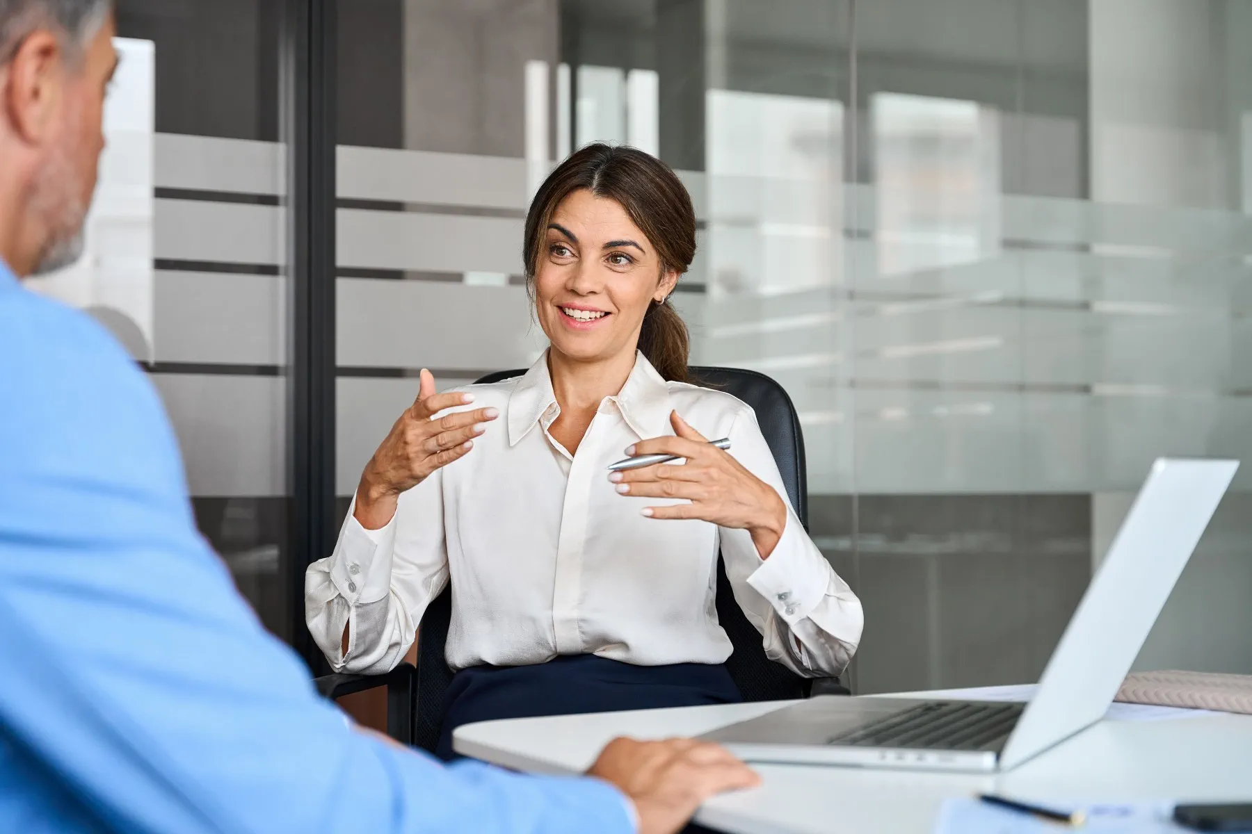 A woman in business attire sits at a desk, speaking and gesturing with her hands to a man across from her in an office setting with a laptop open in front of her.