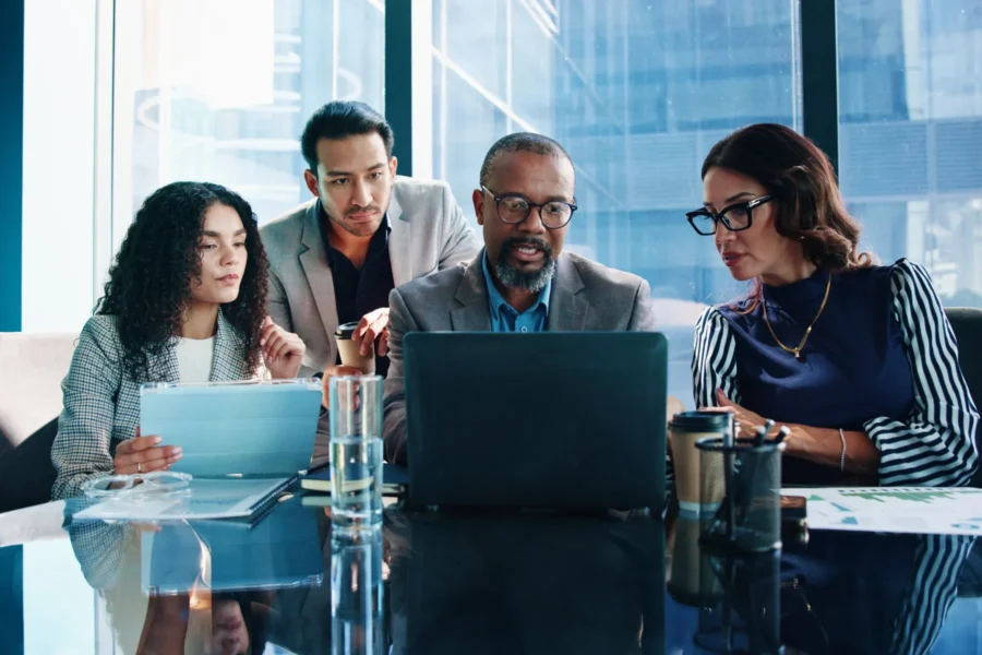 Four people in business attire sit at a table in an office, focused on a laptop screen, with documents, coffee cups, and a glass of water on the table.