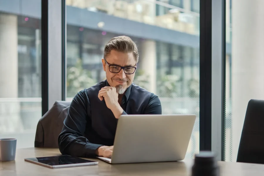 A man with glasses and a goatee sits at a desk, looking at a laptop screen with a slight smile. A tablet, phone, and coffee mug are also on the desk.
