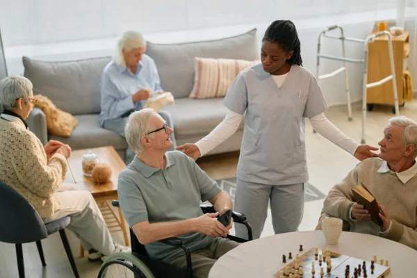 A healthcare worker in scrubs speaks with two senior men seated at a table with a chessboard, as others relax in the background.