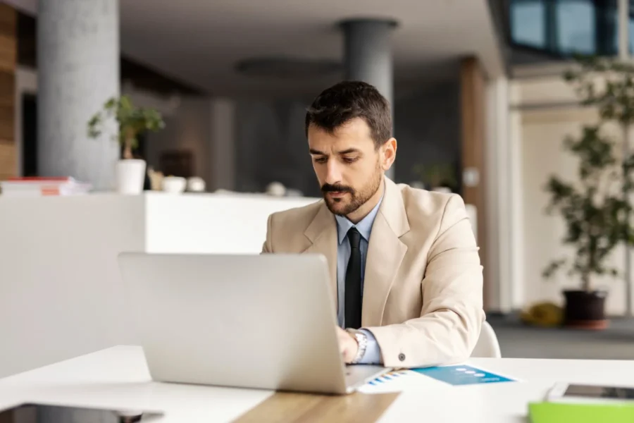 A man in a beige suit works on a laptop at a desk in a modern office setting.