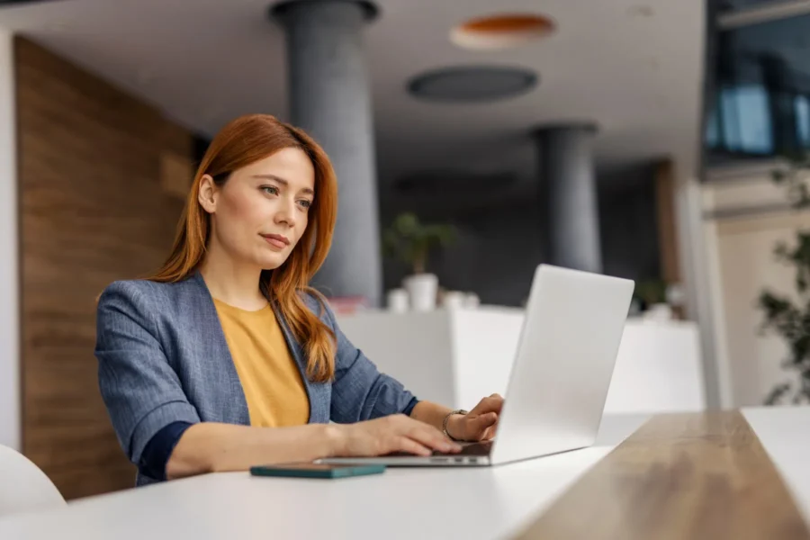 A woman with red hair and a blazer works on a laptop at a modern office desk, with a smartphone beside her.