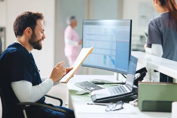 A male healthcare worker sits at a desk reviewing documents, with a computer displaying charts and graphs; two other staff members are visible in the background.