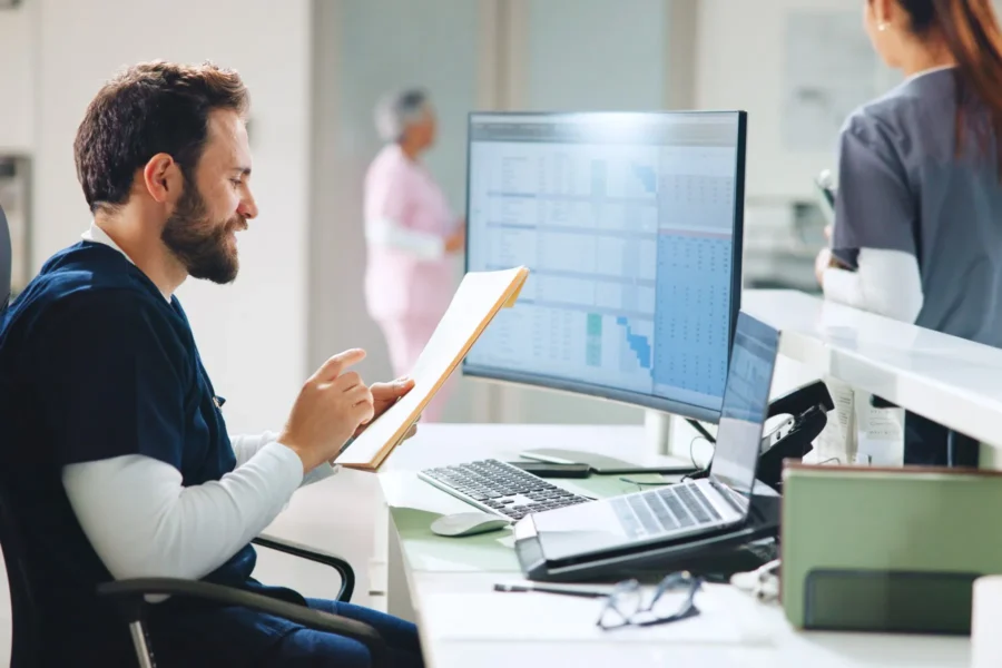 A male healthcare worker sits at a desk reviewing documents, with a computer displaying charts and graphs; two other staff members are visible in the background.