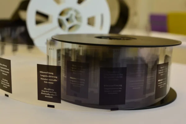 A close-up of a reel of microfilm with visible frames containing text, displayed on a white surface, with another reel blurred in the background.