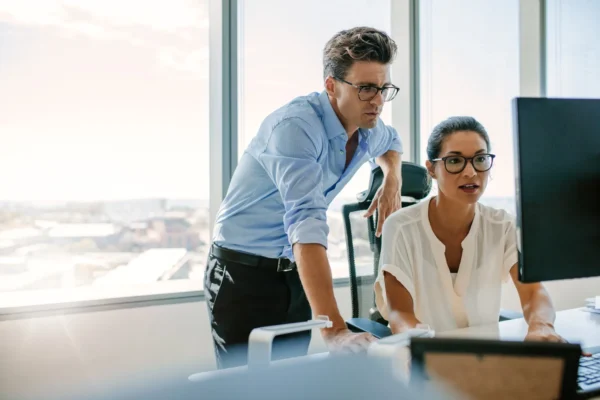 Two colleagues in an office look at a computer monitor together, with one seated and the other standing beside her. Bright windows show a cityscape in the background.