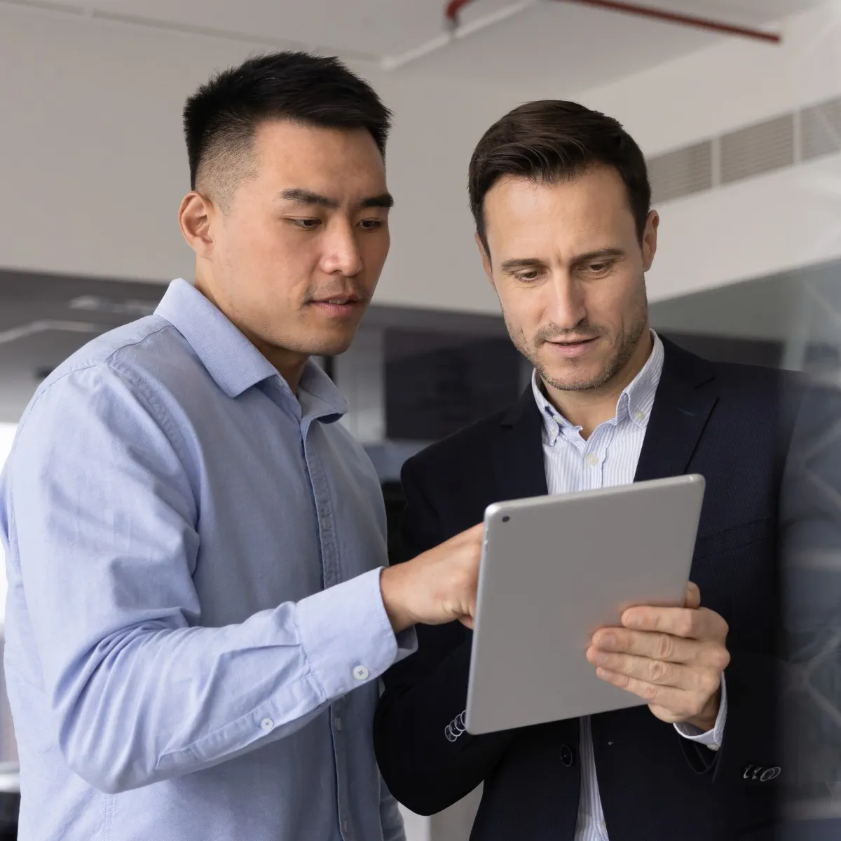 Two men standing indoors, one in a light blue shirt and the other in a dark blazer, looking at and discussing something on a tablet.