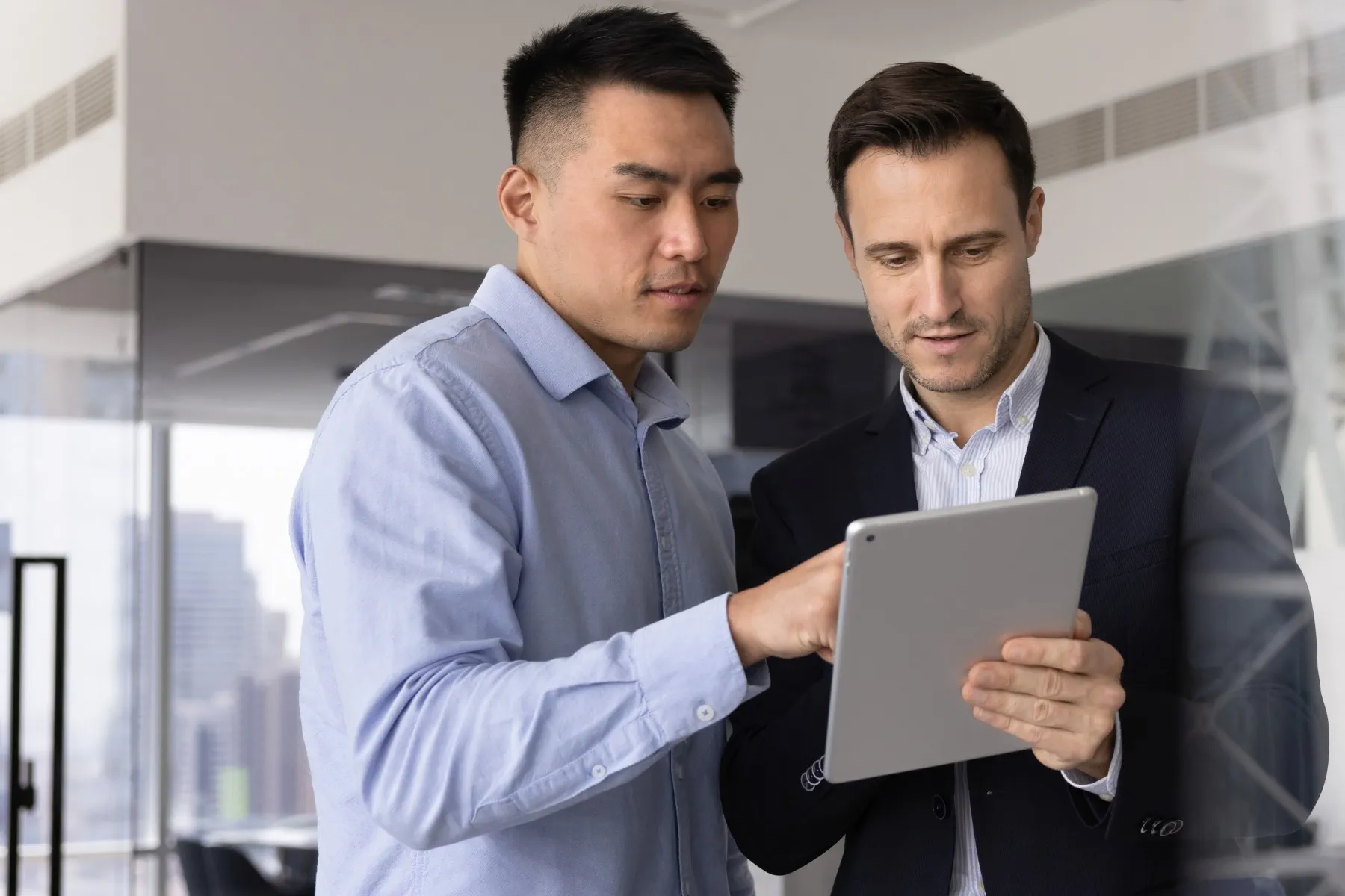 Two men in business attire stand in an office, looking at and discussing something on a tablet device.