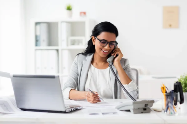 Woman wearing glasses sits at a desk with a laptop, talking on a landline phone and writing on papers in an office setting.