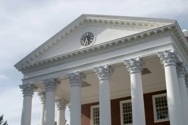 White Greek-style building facade with six Corinthian columns, triangular pediment, and a black and gold clock at the center. Brick wall and windows are visible below the structure.