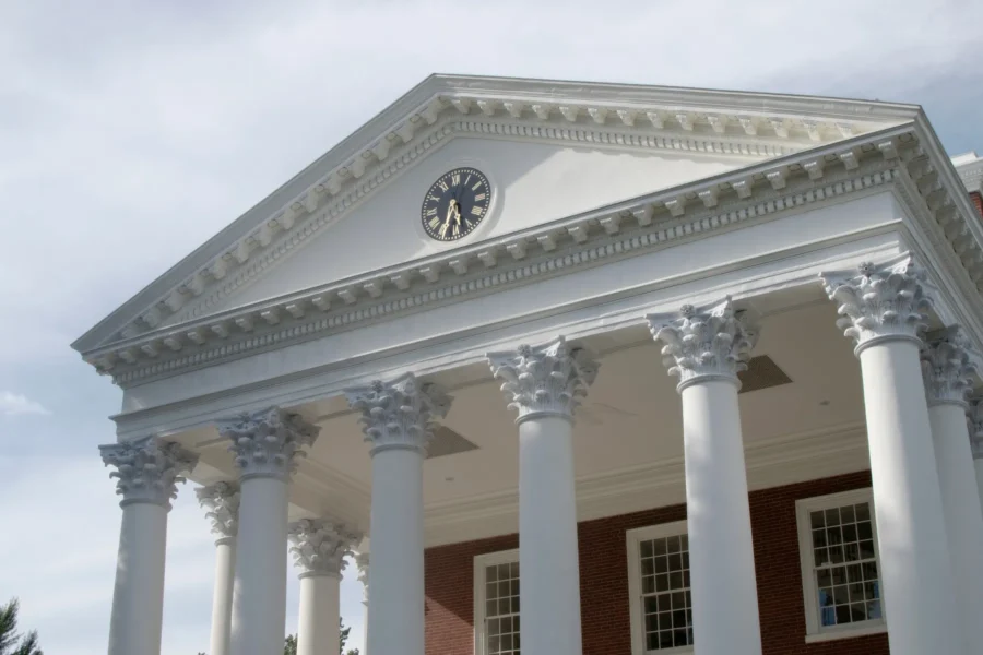 White Greek-style building facade with six Corinthian columns, triangular pediment, and a black and gold clock at the center. Brick wall and windows are visible below the structure.