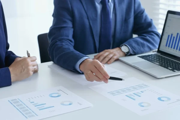 Two people in business attire sit at a table reviewing printed charts and graphs, with a laptop displaying bar graphs nearby.