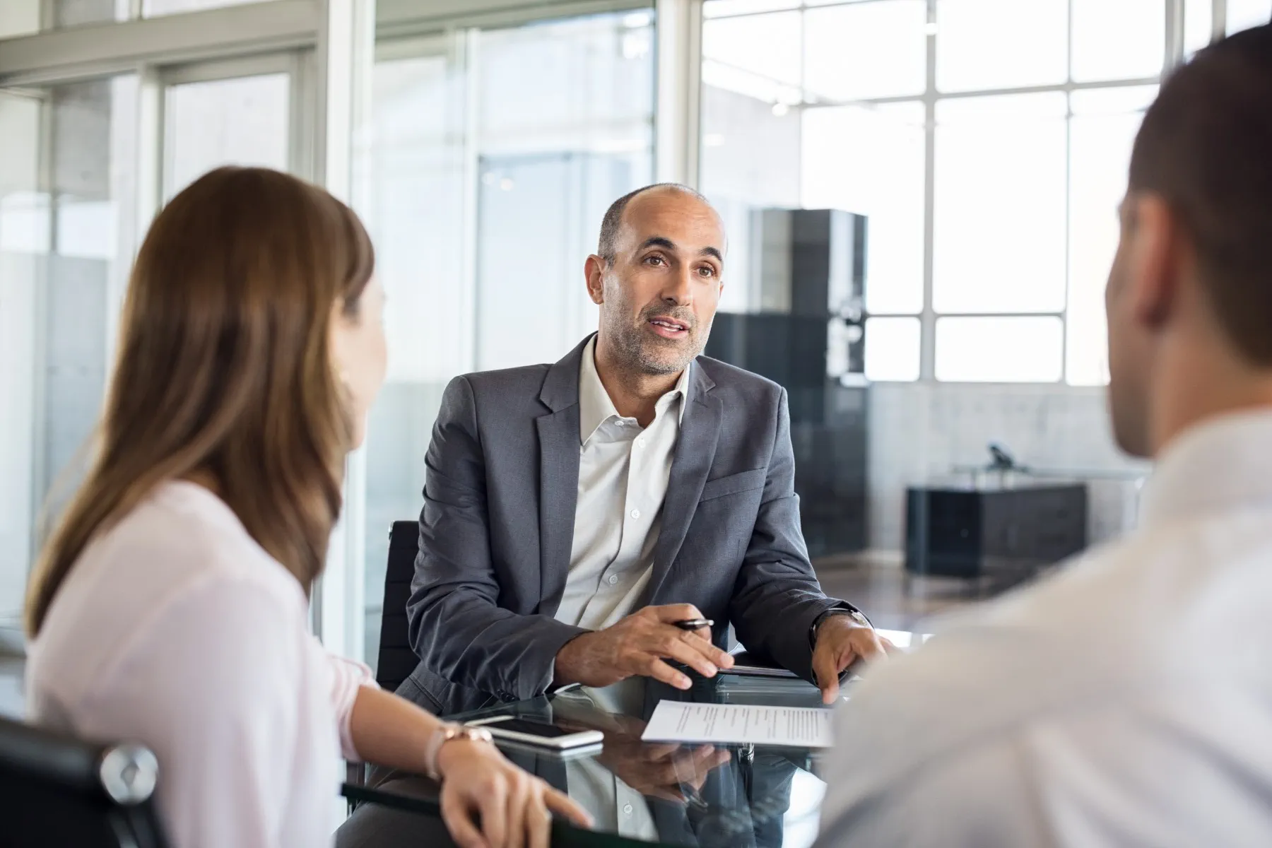 Three people in business attire are sitting at a glass table in an office, having a discussion with documents in front of them.