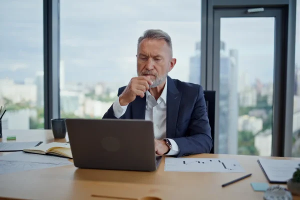 An older man in a suit sits at a desk in a modern office, looking thoughtfully at a laptop, with documents, a notebook, and a cup on the table.