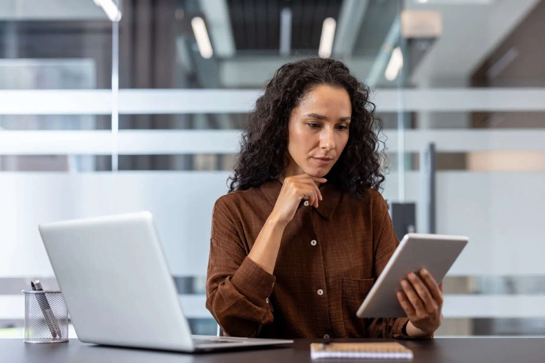 A woman sits at a desk with a laptop, looking thoughtfully at a tablet. A notepad and pen are on the desk in front of her. The office background features glass partitions.