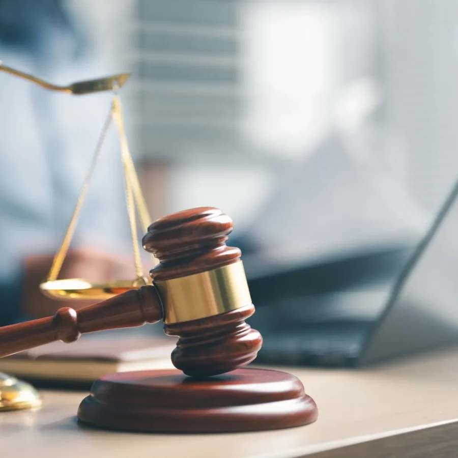 A wooden judge’s gavel rests on its sound block with a brass balance scale and an open laptop in the background.