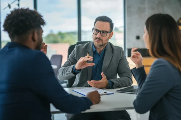 Three people in business attire sit at a table in an office, engaged in a serious discussion; one man gestures with his hand while speaking.