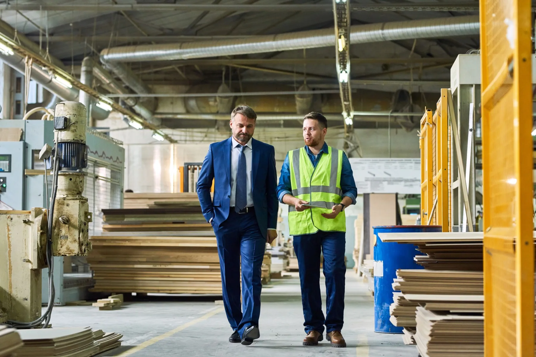 A man in a suit walks with a man in a reflective vest inside a warehouse with stacks of wood and industrial equipment.