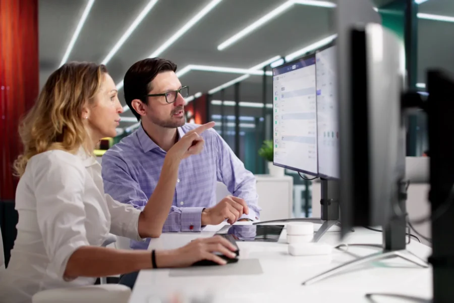 Two colleagues sit at a desk in a modern office, looking at a large computer monitor and discussing information displayed on the screen.