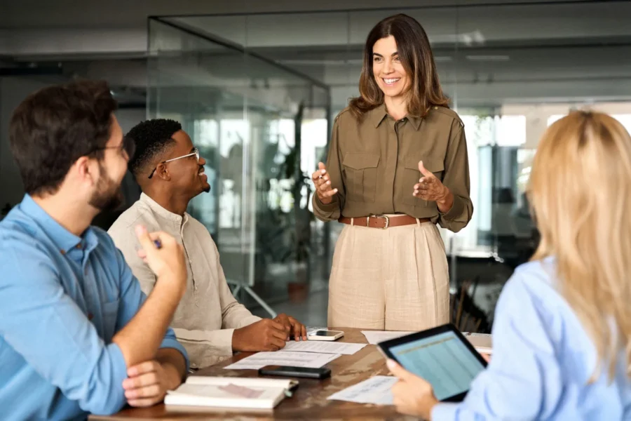 Four people in business attire have a meeting around a table in a modern office, with one woman standing and speaking while the others listen and take notes.