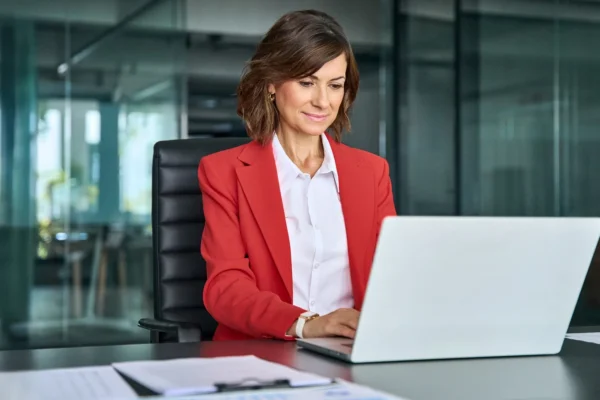 A woman in a red blazer sits at a desk working on a laptop in a modern office setting.
